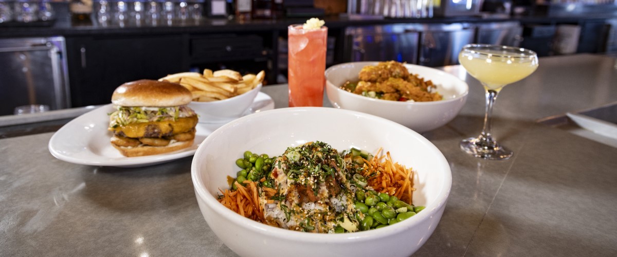  Cobb Chicken Salad, Pickle Back Burger with Fries, and Spicy Tuna Bowl in white dishes along with Cherry Blossom Limeade in a tall glass, garnished with a flower and Passionfruit Daquiri on the bar counter of Sports and Social with the bar in the background.